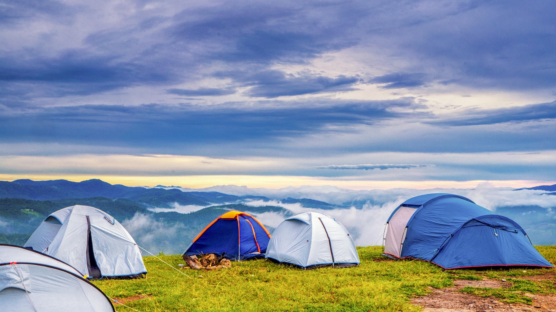 Mehrere Zelte stehen auf einer grünen Wiese oberhalb der Wolken, mit Blick auf ein Bergpanorama bei bewölktem Himmel.